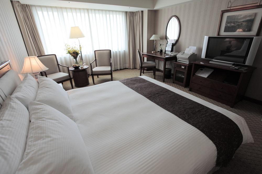 فندق إيفرجرين لوريل، تايتشونج تايتشونغ المظهر الخارجي الصورة The photo shows a well-furnished hotel room. In the foreground, there is a large bed with white bedding and a dark-colored throw at the foot. Beside the bed, there is a bedside lamp. 

In the background, natural light filters through large windows co