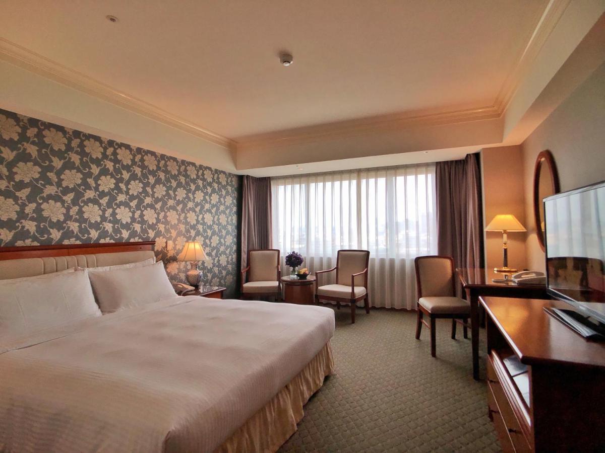 فندق إيفرجرين لوريل، تايتشونج تايتشونغ المظهر الخارجي الصورة The photo shows a well-appointed hotel room. It features a large bed with white bedding and a decorative headboard. The walls are adorned with a patterned wallpaper, possibly floral, adding a touch of elegance. There is a window with sheer curtains t