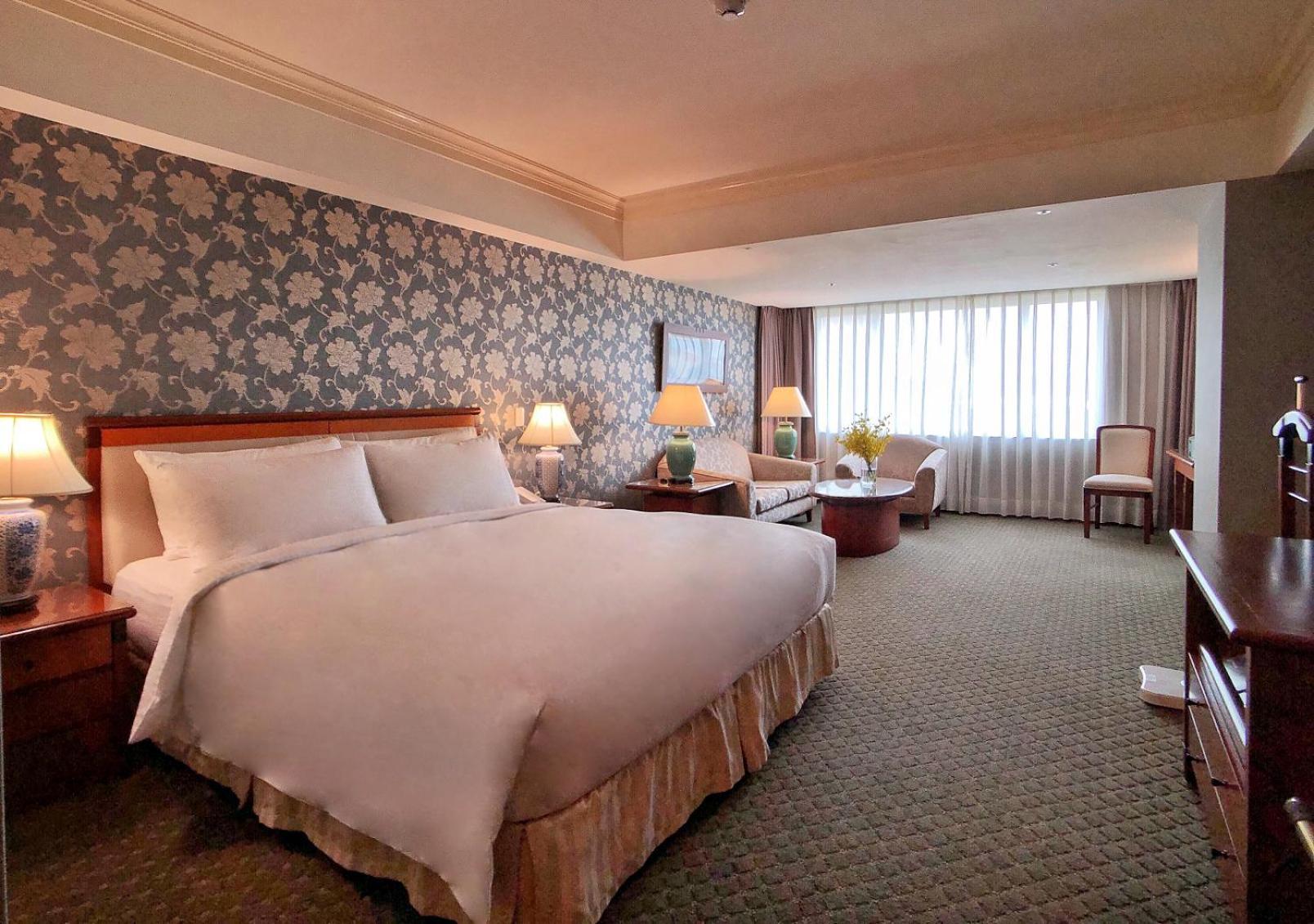 فندق إيفرجرين لوريل، تايتشونج تايتشونغ المظهر الخارجي الصورة The photo shows a hotel room with a spacious and comfortable layout. It features a large bed with white bedding, placed against a decorative wallpapered wall. On either side of the bed, there are lamps providing soft lighting. In addition to the bed,