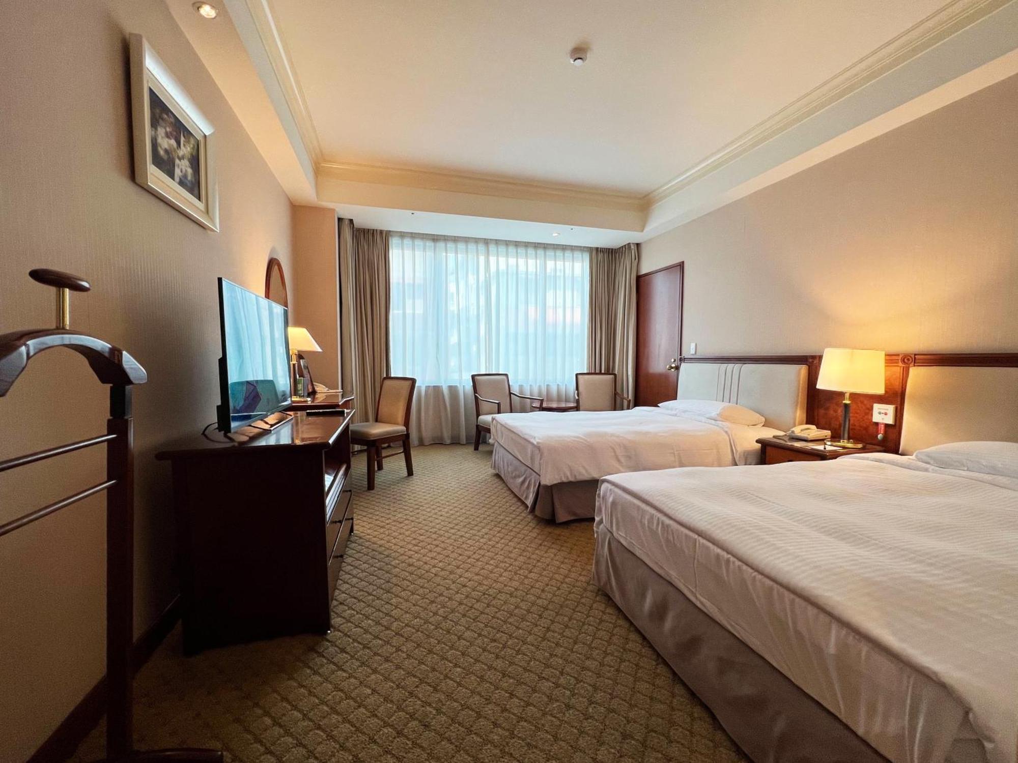 فندق إيفرجرين لوريل، تايتشونج تايتشونغ المظهر الخارجي الصورة The photo shows a hotel room featuring two twin beds placed parallel to each other. The room has a warm color palette and includes a large window with sheer curtains allowing natural light to illuminate the space. There is a wooden desk and two chair