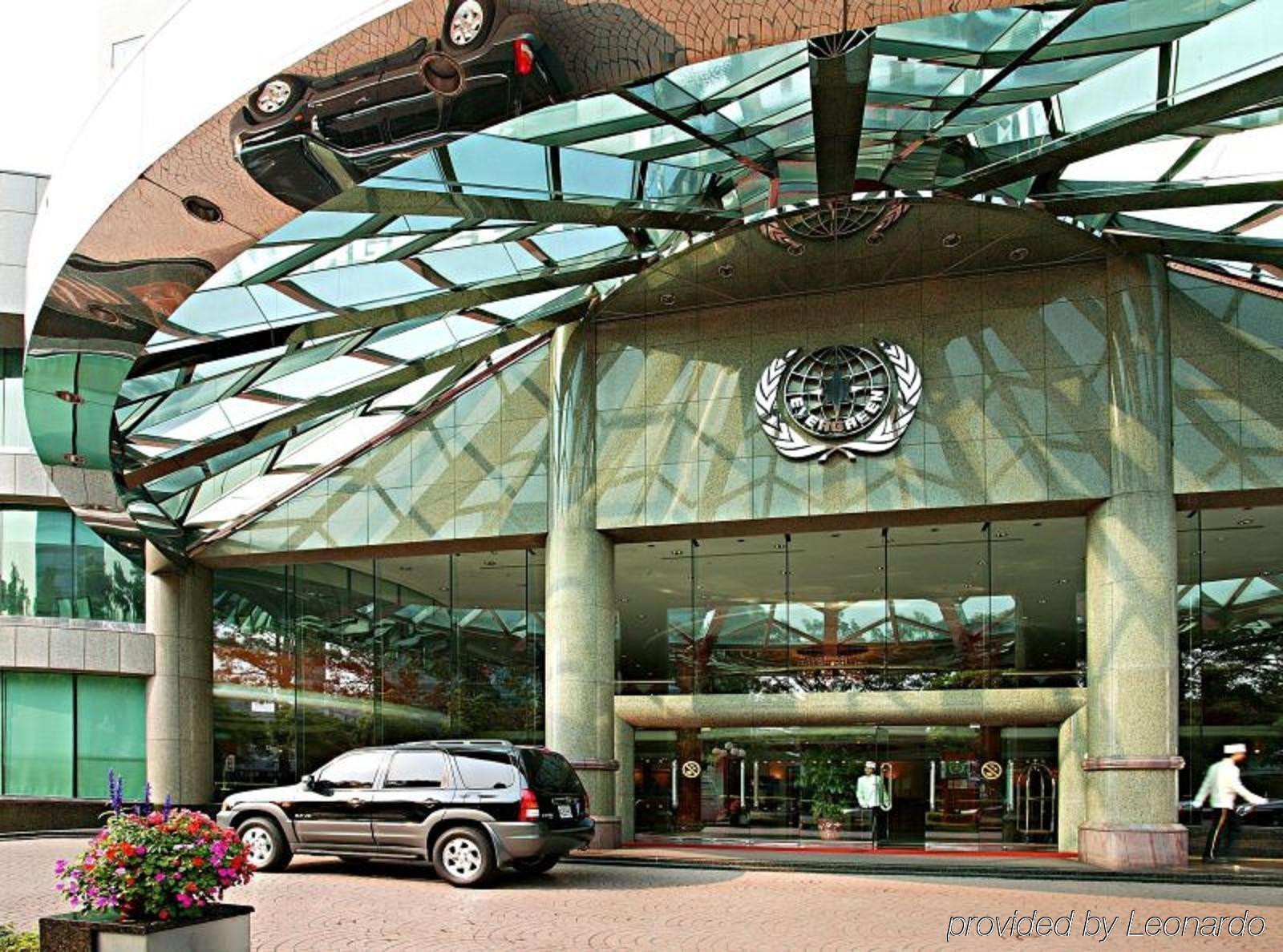 فندق إيفرجرين لوريل، تايتشونج تايتشونغ المظهر الخارجي الصورة The photo shows the entrance of a modern building characterized by its glass and metal architecture. The structure features a large, domed roof that reflects light, with a distinctive emblem or logo displayed prominently at the center. In front of th