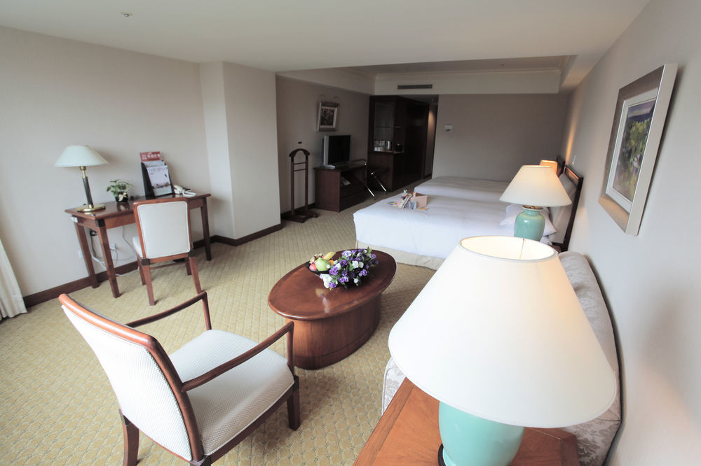 فندق إيفرجرين لوريل، تايتشونج تايتشونغ المظهر الخارجي الصورة The photo shows an interior view of a hotel room. It features two beds, a coffee table with a small decorative arrangement of flowers, and a comfortable chair. There's a desk and a lamp, along with another larger lamp on the table. In the background,