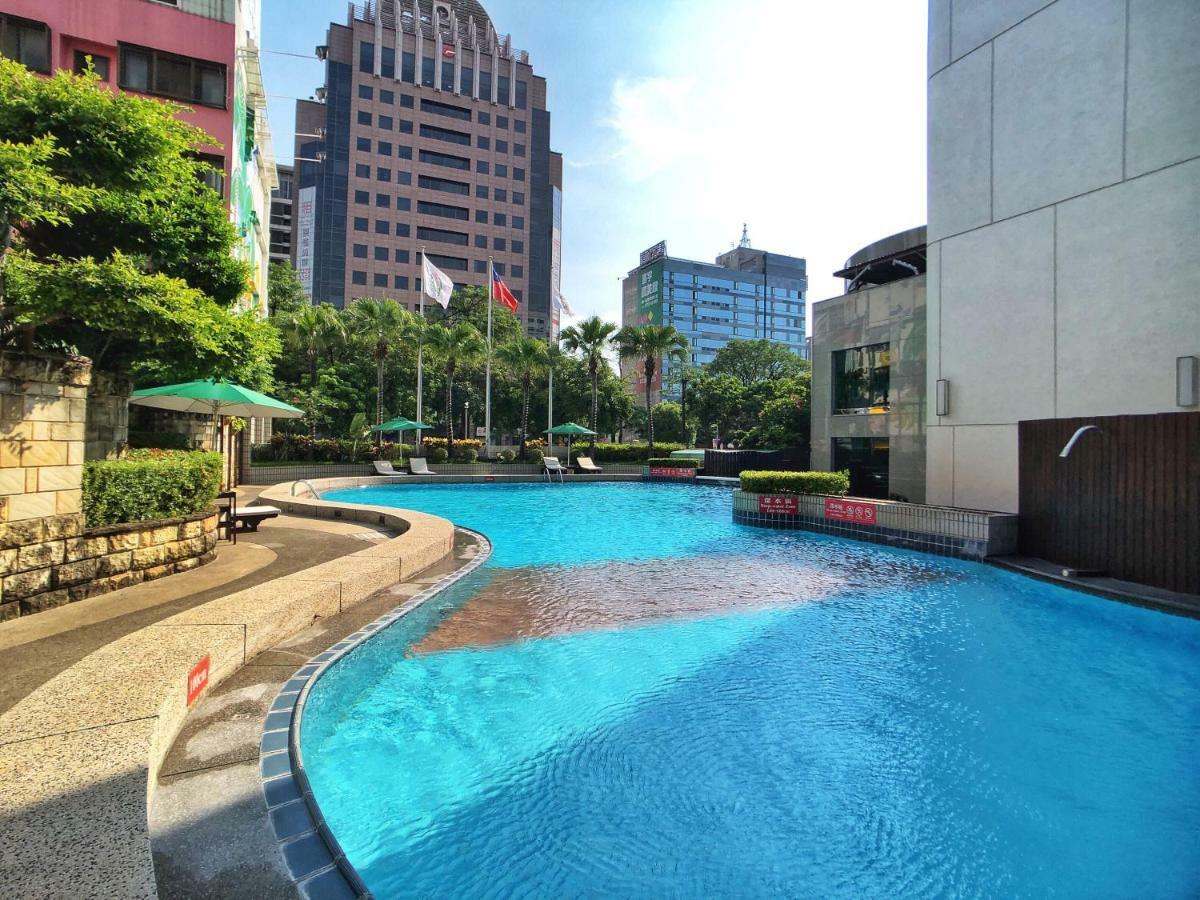 فندق إيفرجرين لوريل، تايتشونج تايتشونغ المظهر الخارجي الصورة The photo shows a swimming pool set in a modern urban environment. The pool has a curved edge and is surrounded by greenery, including trees and umbrellas for shade. In the background, there are several tall buildings, indicating a cityscape, with so