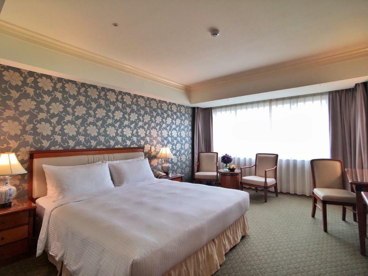 فندق إيفرجرين لوريل، تايتشونج تايتشونغ المظهر الخارجي الصورة The photo shows a hotel room that features a large bed with a white bedspread positioned against a decorative floral wallpaper. There are two bedside lamps on wooden nightstands flanking the bed. To the side of the room, there are two chairs and a sm