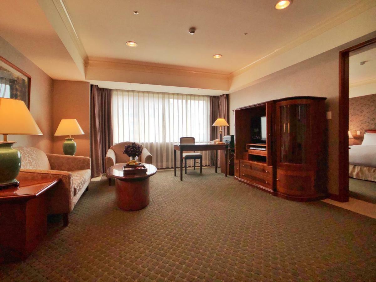 فندق إيفرجرين لوريل، تايتشونج تايتشونغ المظهر الخارجي الصورة The photo shows a cozy hotel suite or living area. There is a sofa and an armchair arranged around a small coffee table. A desk with a chair is positioned near a large window covered with sheer curtains that allow natural light to enter the room. Two