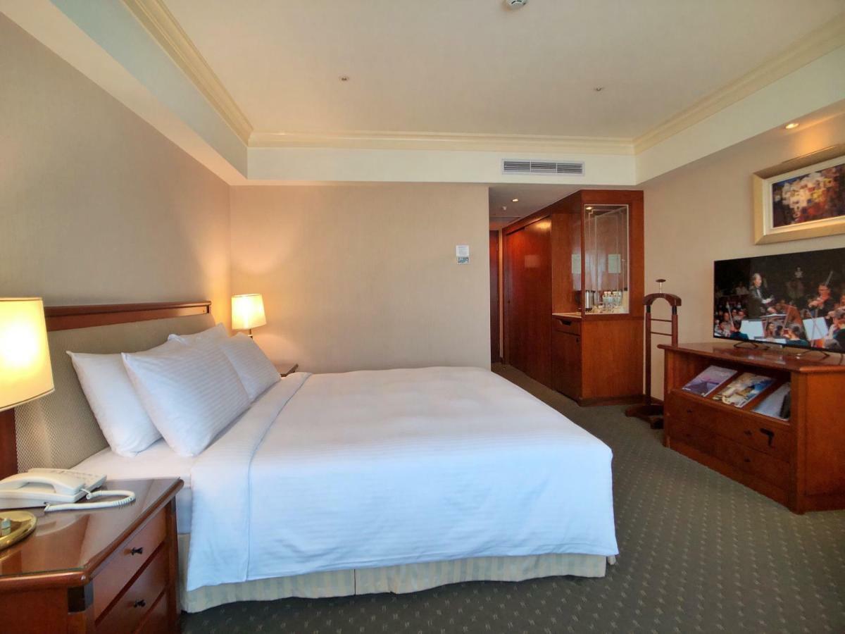 فندق إيفرجرين لوريل، تايتشونج تايتشونغ المظهر الخارجي الصورة The photo shows a hotel room featuring a comfortable bed with white linens and pillows, positioned against a light-colored wall. There is a wooden bedside table with a lamp and an open book next to the bed. On the opposite side, there’s a large flat-