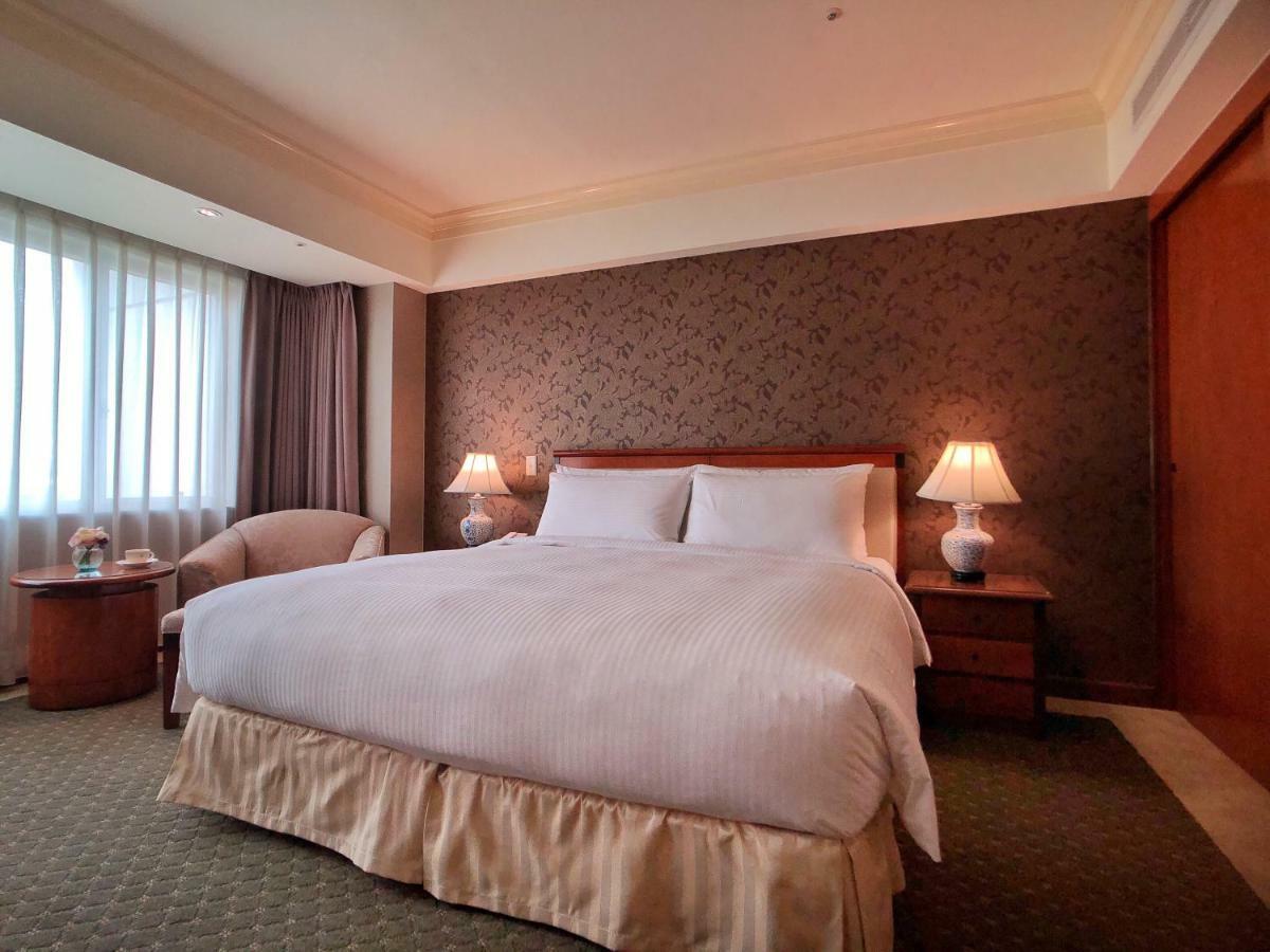 فندق إيفرجرين لوريل، تايتشونج تايتشونغ المظهر الخارجي الصورة The photo shows a neatly arranged hotel room featuring a large bed with white linens and a light-colored bed skirt. On either side of the bed, there are small wooden nightstands with lamps. To the left of the bed, there's a comfortable chair and a ro
