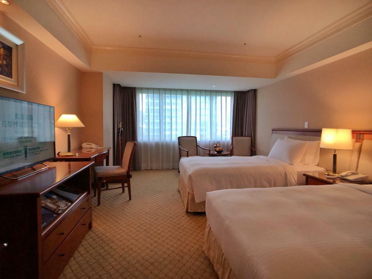 فندق إيفرجرين لوريل، تايتشونج تايتشونغ المظهر الخارجي الصورة The photo shows a hotel room featuring two twin beds with white bedding. There's a television mounted on a wooden dresser, and next to it, a comfortable chair and a small table are positioned. The room has large windows that allow natural light to en