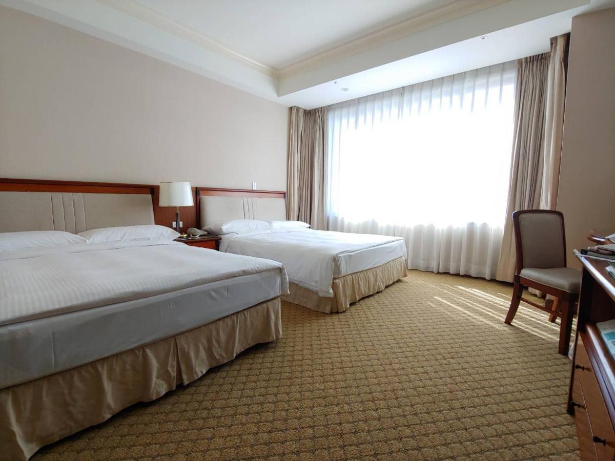 فندق إيفرجرين لوريل، تايتشونج تايتشونغ المظهر الخارجي الصورة The photo shows a neatly arranged hotel room featuring two beds: one double bed and one single bed. The room has beige walls and a warm color palette. Large windows with sheer curtains allow natural light to flood in, creating a bright and airy atmos