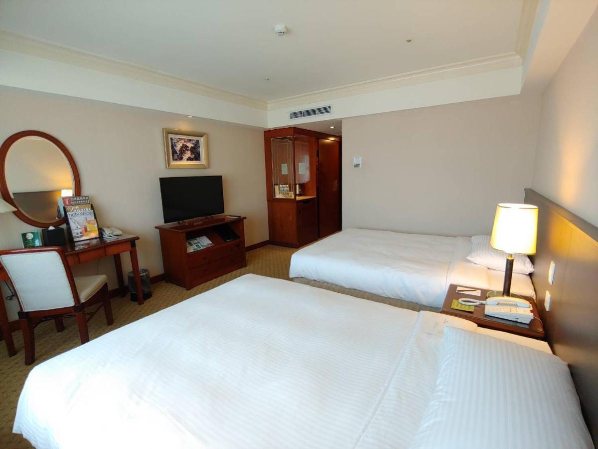 فندق إيفرجرين لوريل، تايتشونج تايتشونغ المظهر الخارجي الصورة The photo shows a hotel room with two beds, neatly made with white linens. There is a desk and chair in one corner, accompanied by a flat-screen TV mounted on the wall. A large mirror and a wooden cabinet are also visible. The room is illuminated by 